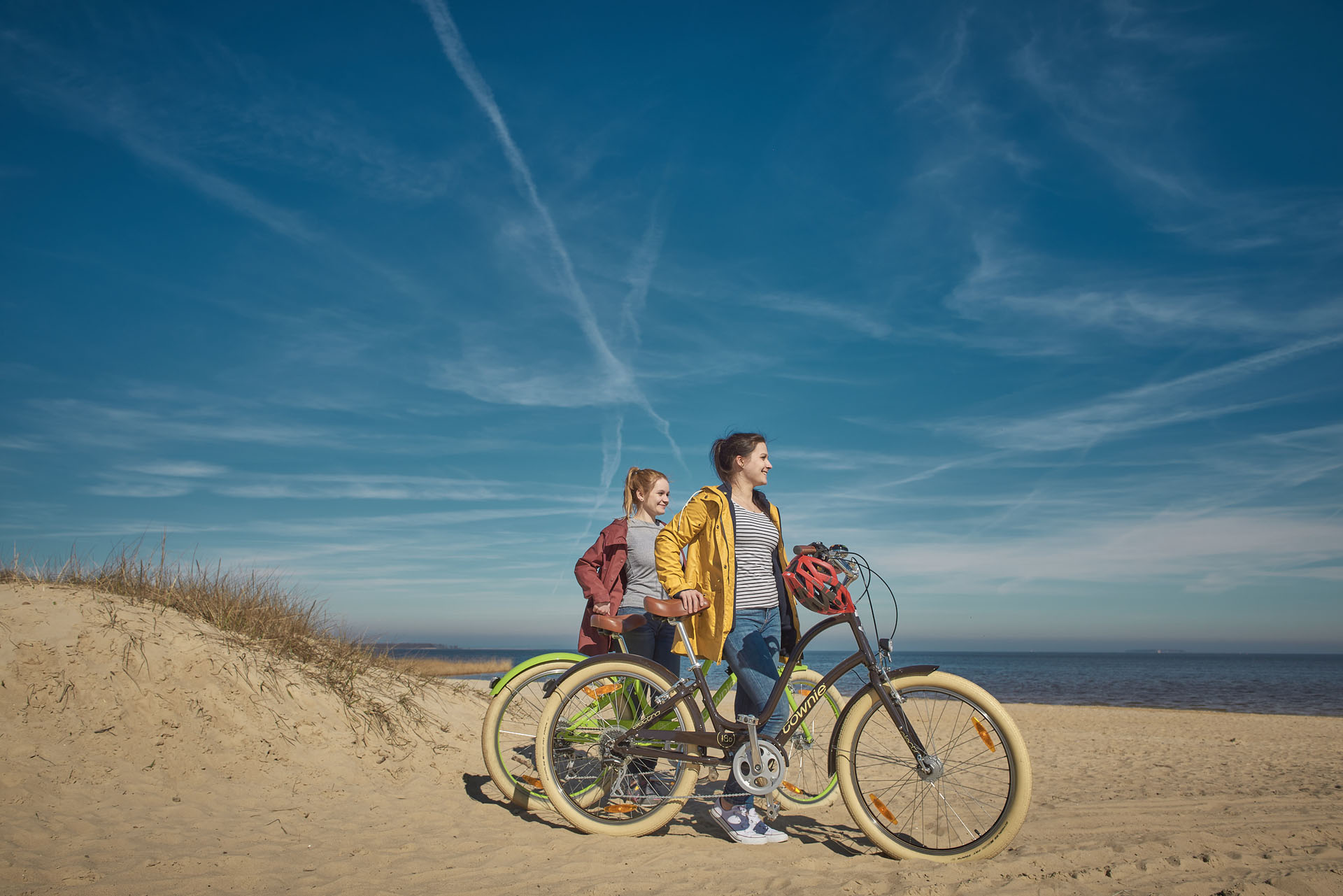 There are two women on the beach who push their bikes through the sand. On the left is a small dune and the water in the background.